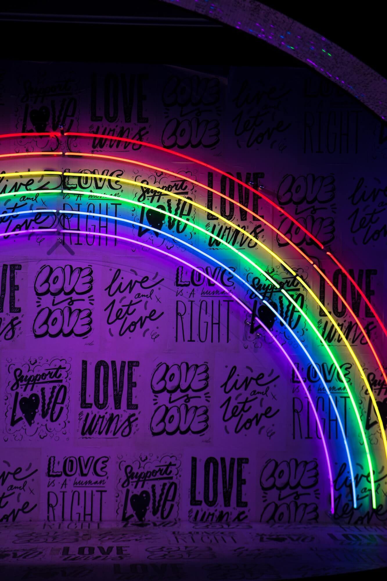 Neon rainbow sign against a background of "Love is Love" and "Love Wins" wallpaper