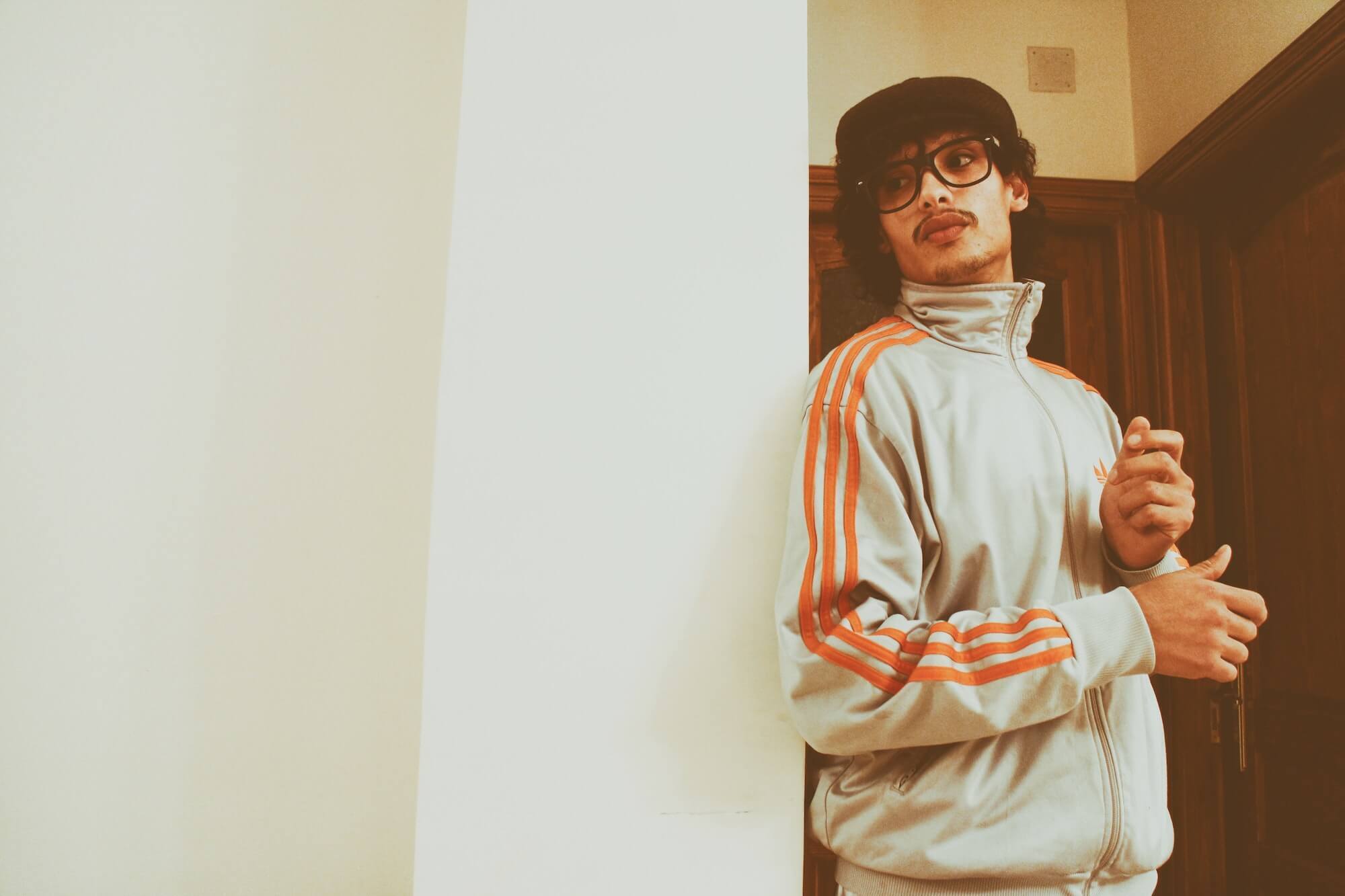 Man in hat, sunglasses and Adidas Three-Stripes track jacket leans against wall