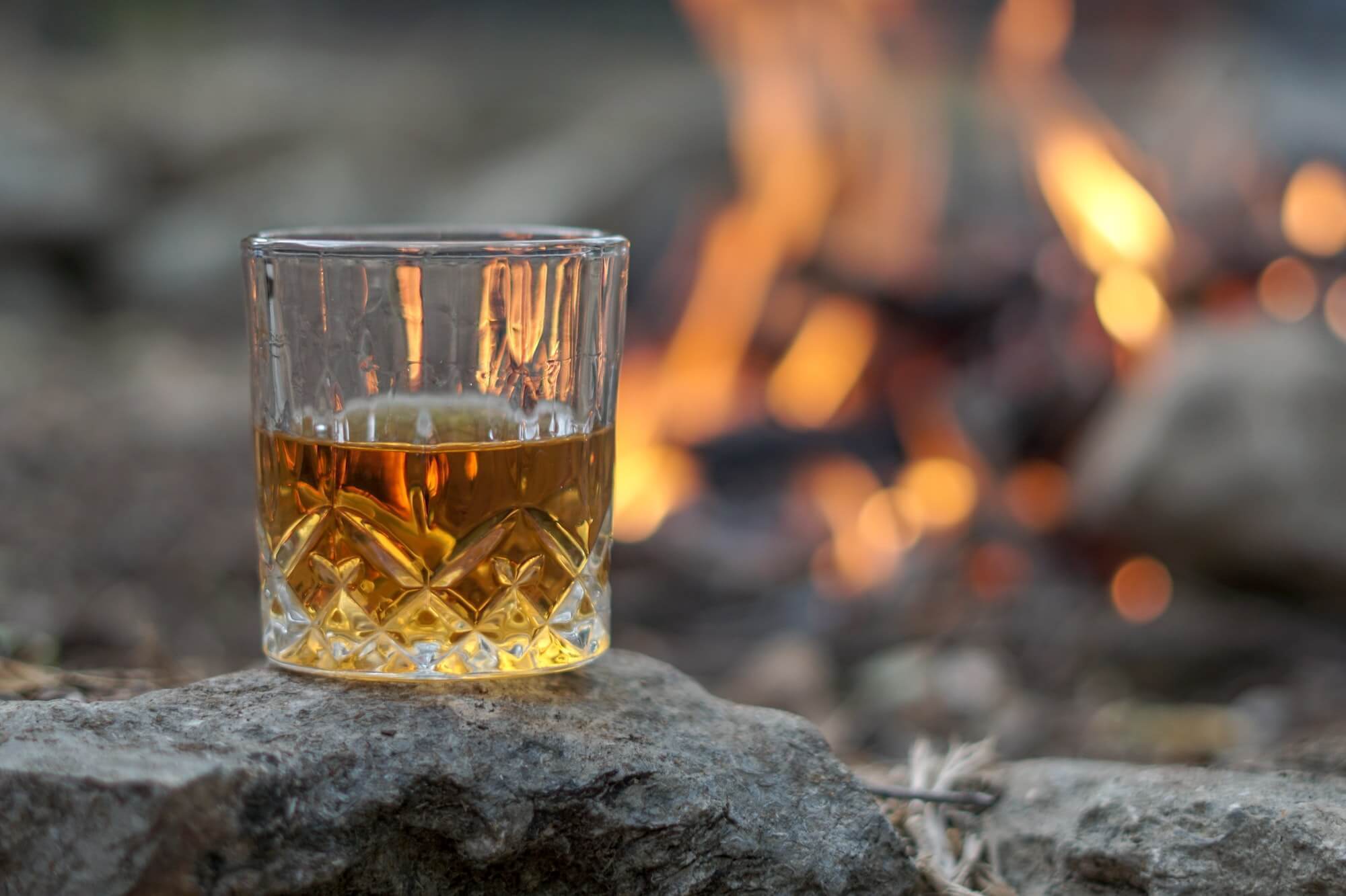 Crystal tumbler with whiskey atop a rock with campfire in the background