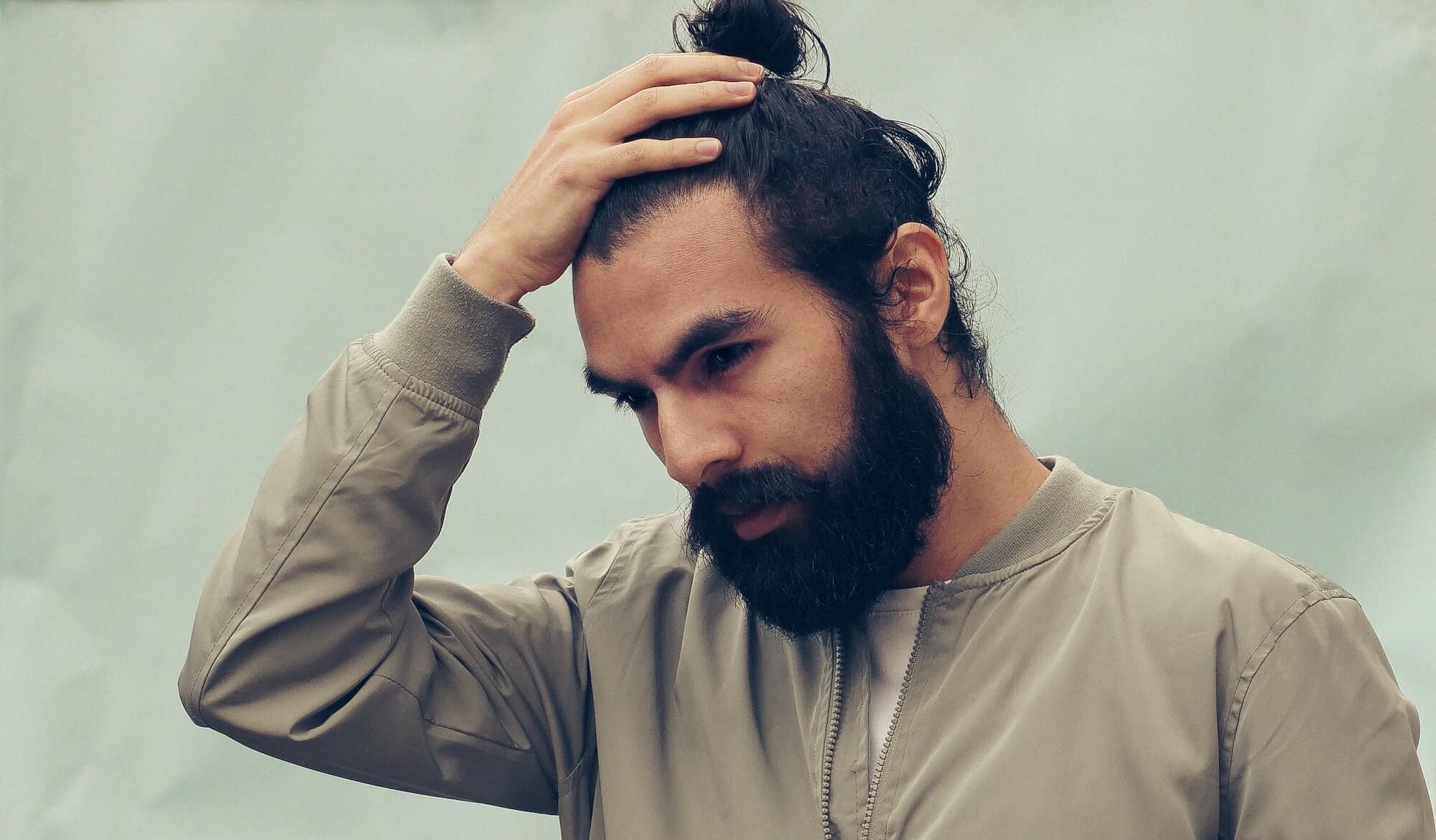 Black-haired man with beard, mustache and man bun holds his hand up to his hairline