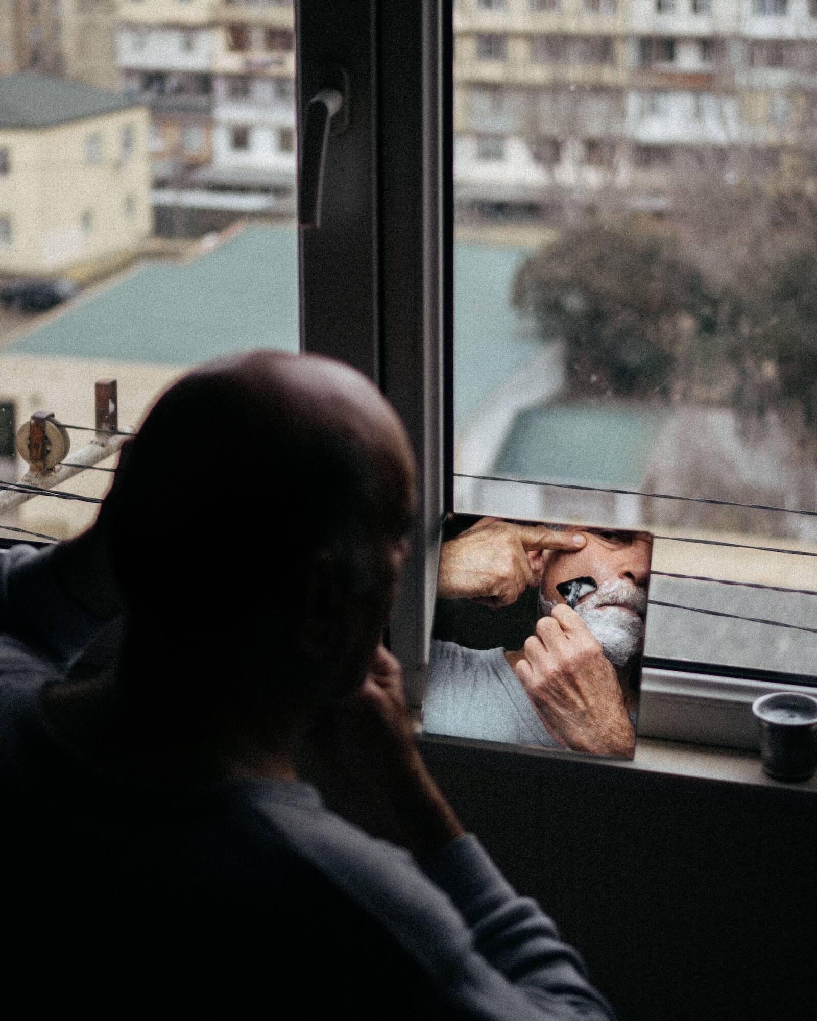 Man shaving his face in a small mirror in front of window