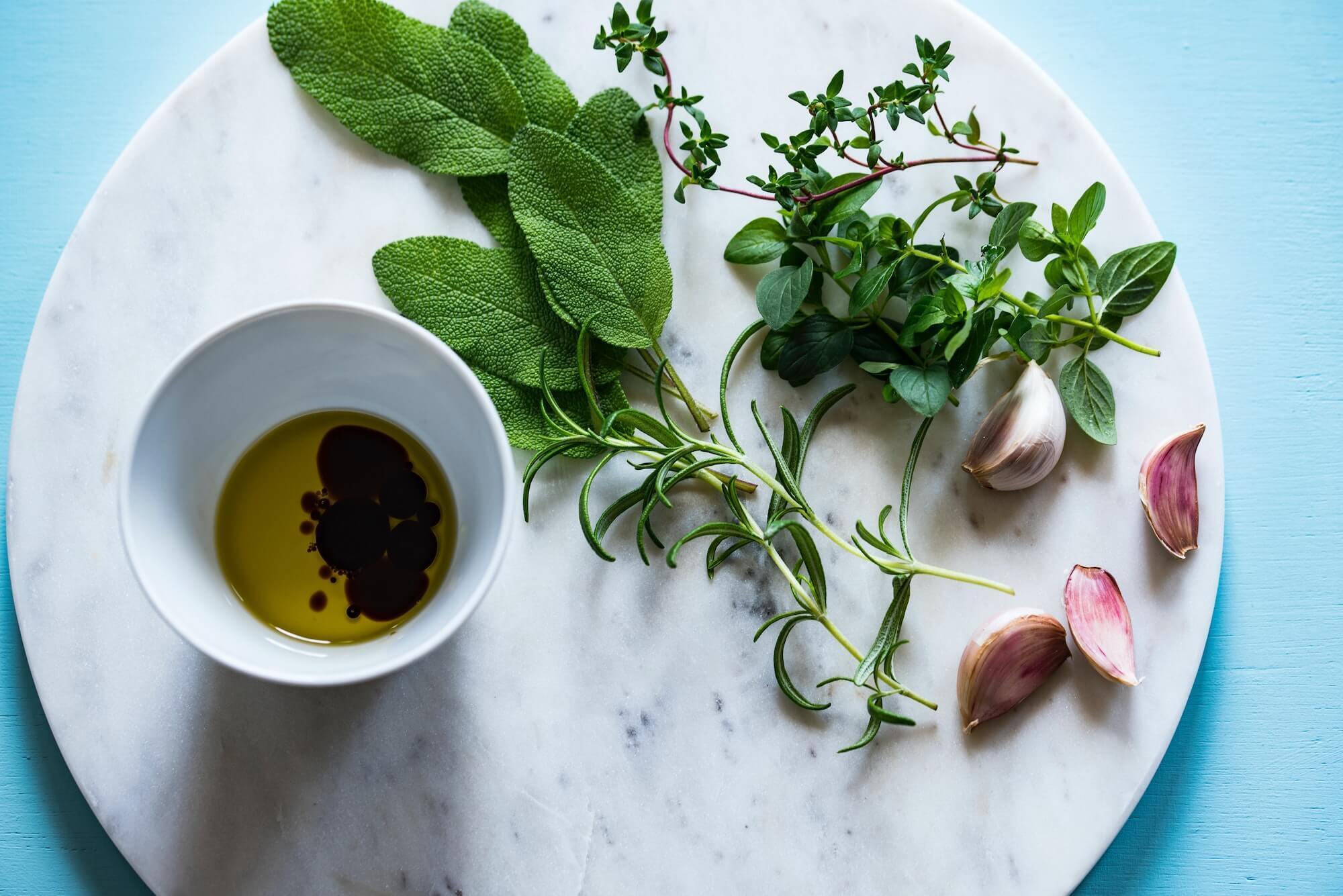 Cup with olive oil and balsamic vinegar next to fresh herbs and garlic