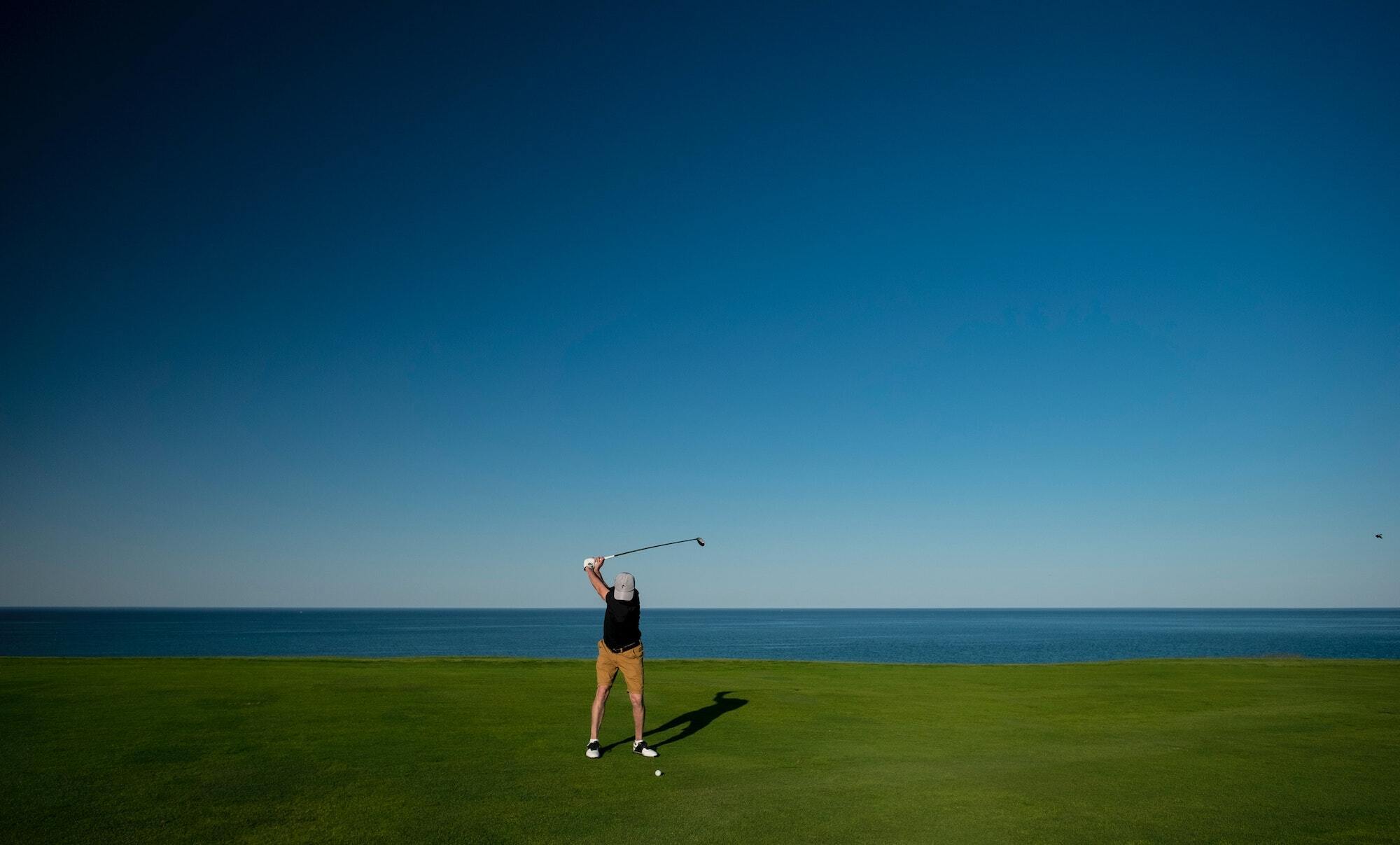 Man swinging golf club with blue sky and ocean in background