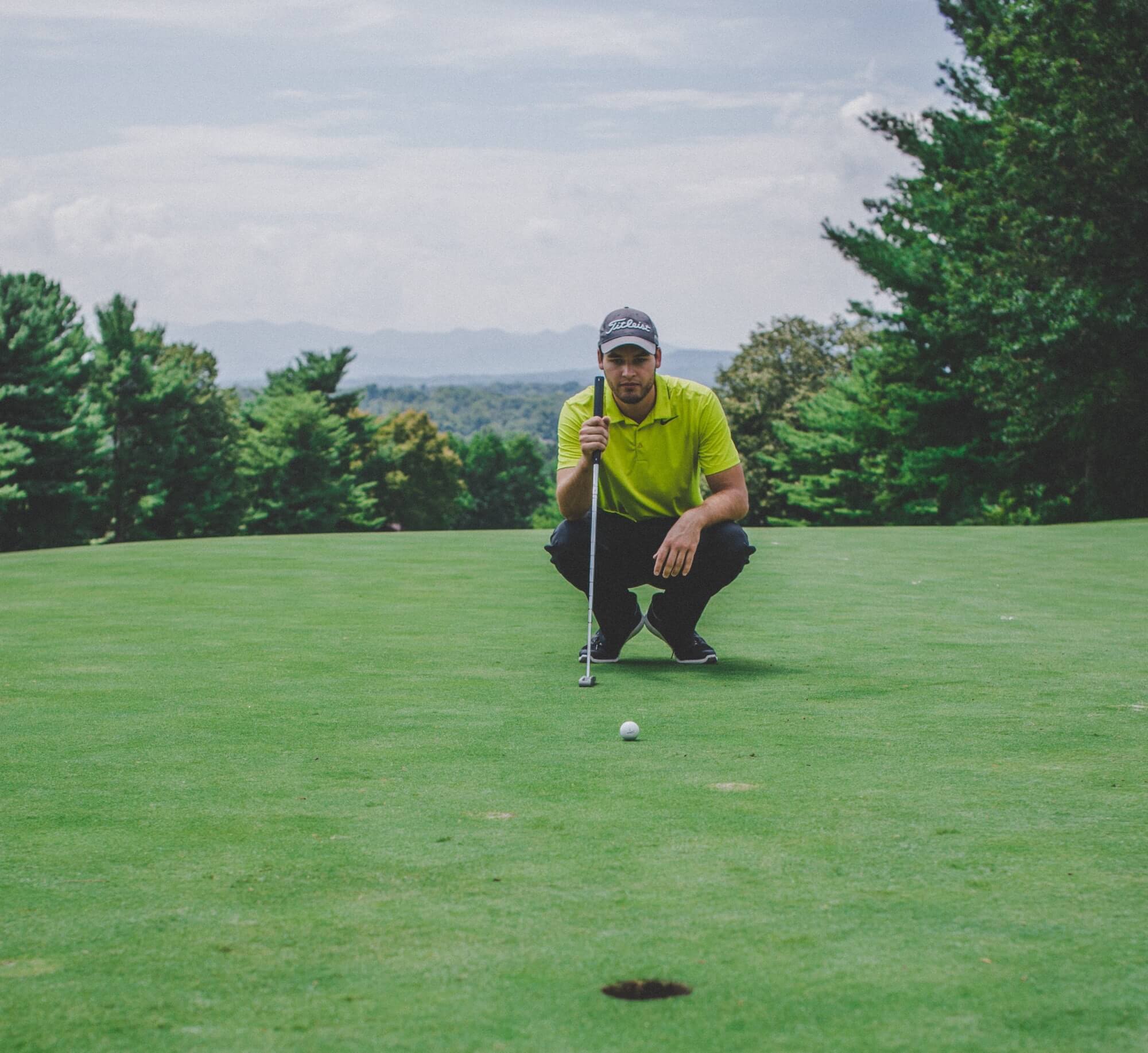 Man in Titleist cap and Nike golf shirt crouches by cup on golf coarse