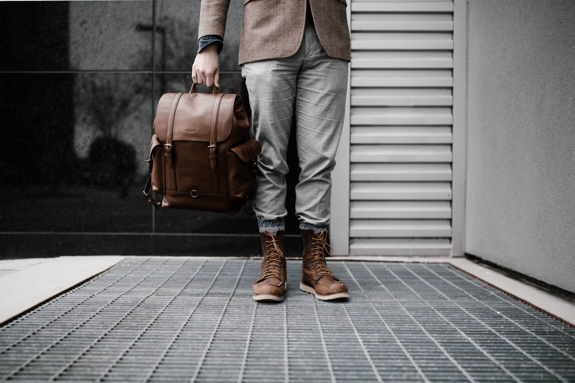 Man in brown blazer and brown leather lace-up boots holds brown leather backpack in his right hand