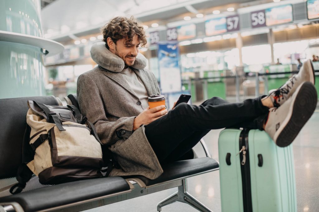 Man in airport with travel pillow around neck and legs up on luggage holds coffee and looks at phone